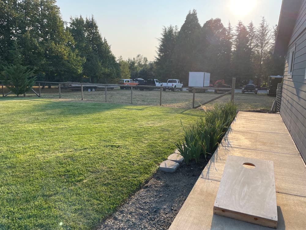 Backyard view with green lawn, cornhole board, trees, and parked vehicles in the background.