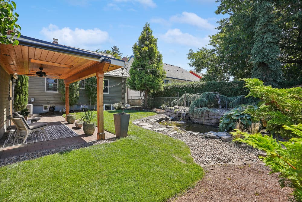 Lush backyard with a pond, stone path, and covered seating area amidst greenery.