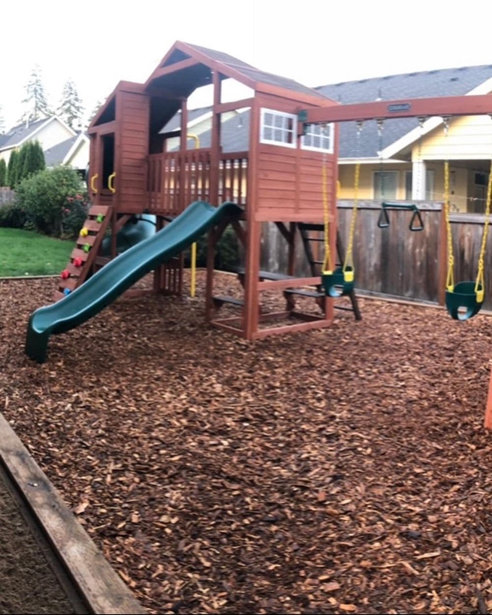 Wooden playset with slide, swings, and climbing features in a backyard with mulch ground.