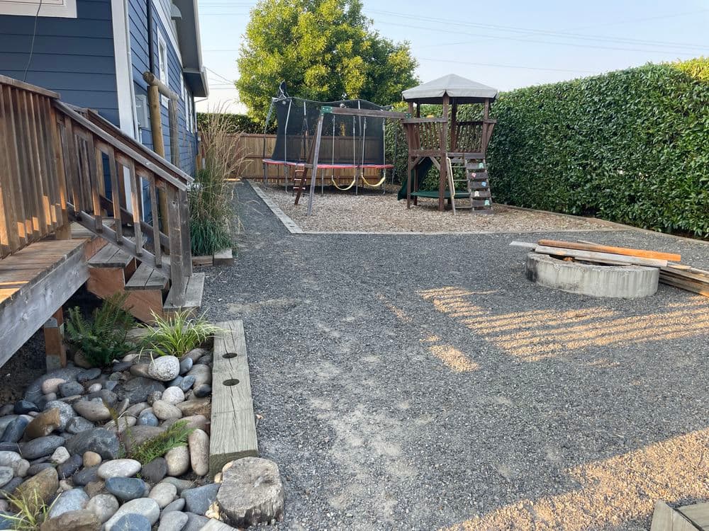 Residential backyard with gravel area, playset, trampoline, and gazebo surrounded by greenery.