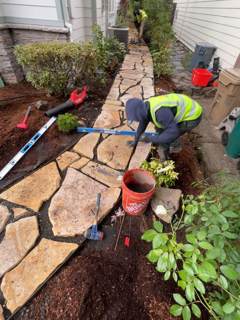 Worker landscaping a stone pathway with tools, plants, and mulch in a residential yard.